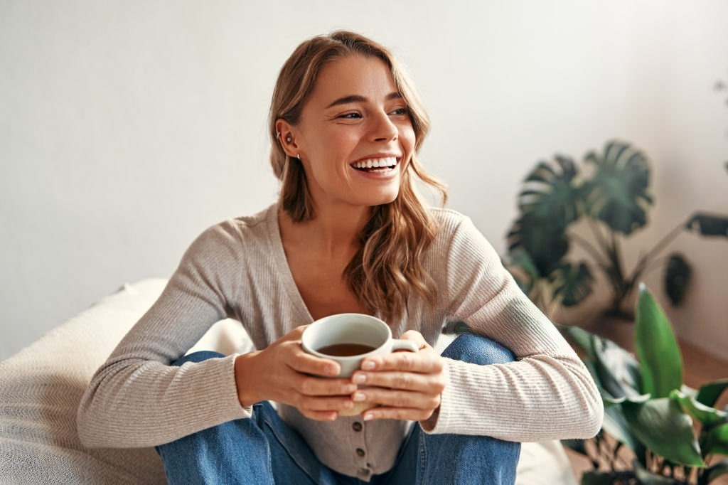 A smiling woman sitting comfortably, holding a cup of tea, with a green plant in the background.